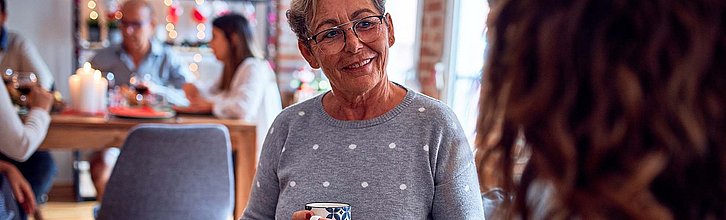 Eine ältere Frau hält eine Tasse in beiden Händen und unterhält sich. Im Hintergrund sitzen mehrere Personen an einem Tisch.